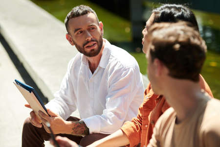 Young businessman in white shirt holding tablet computer when talking to colleagues outdoorsの写真素材