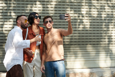 Group of positive friends in sunglasses taking selfie on smartphone when walking in city