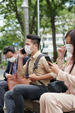 Frowning young Asian man in protective mask talking on phone when sitting on bench outdoors
