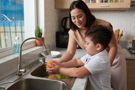 Little boy rinsing fruits under tap water under control on his motherの写真素材