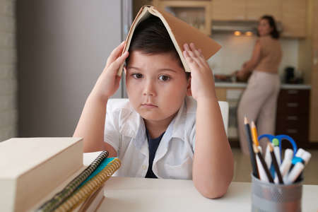 Sad boy covering head with opened book when sitting at kitchen tableの写真素材