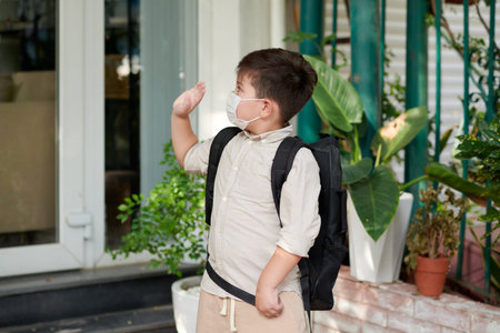 Schoolboy in medical mask waving with hand when leaving house for schoolの写真素材