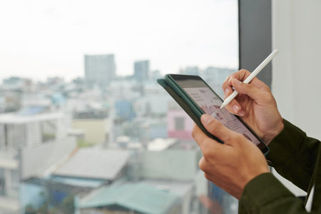 Hands of entrepreneur standing at office window and checking charts on tablet computer