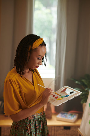 Creative Black woman mixing paints on palette when painting picture on easel