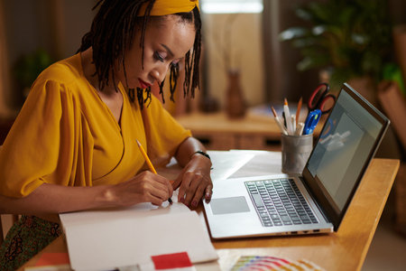 Fashion designer working at desk with opened laptop and drawing sketches in sketchbook