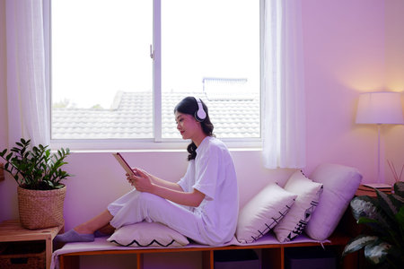 Teenage girl in headphones sitting at window and watching show on tablet computer