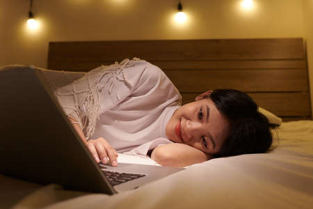 Smiling girl resting on bed and watching romantic comedy movie on laptopの写真素材