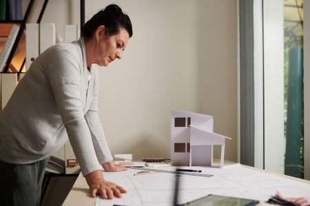 Pensive mature female architect leaning on table with building blueprint and paper house