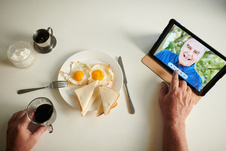 Senior man having fried eggs and coffee when video calling his best friend or brother