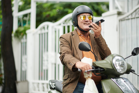 Joyful elderly man sitting on scooter and recording voice message for friend or girlfriend