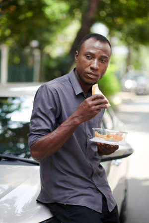 Portrait of taxi driver leaning on car and eating sandwich for lunchの写真素材
