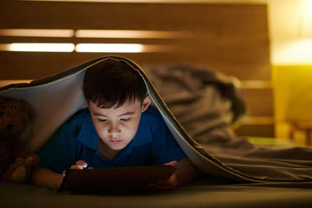 Preteen boy lying under blanket and playing game on tablet computer late at nightの写真素材