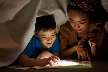 Mother shining flashlight on book page when son reading scary storyの写真素材