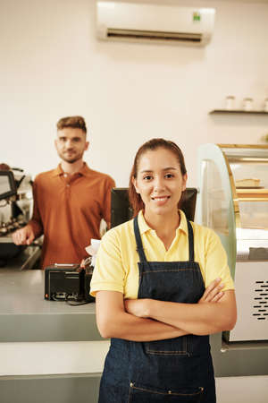 Smiling confident coffeeshop waiter standing with arms crossed, barista working in backgroundの写真素材