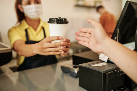 Coffeeshop barista wearing medical mask when selling cup of take out coffee to customerの写真素材