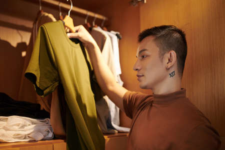 Young man standing in closet and holding hanger with t-short when choosing what to wear todayの写真素材