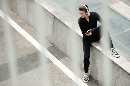 Young female athlete in headphones using smartphone to listen to music while sitting outdoorsの写真素材