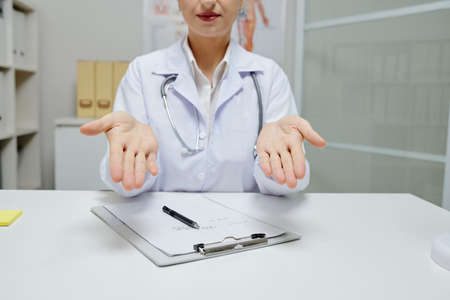 Close-up of female doctor in white coat gesturing while sitting at table with medical card during online meetingの写真素材