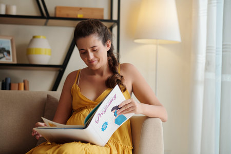 Young pregnant woman sitting on sofa and reading book about pregnancy during her leisure time at...
