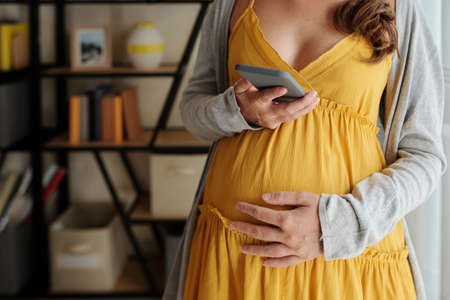 Close-up of pregnant woman stroking her pregnant belly and swiping screen of her smartphoneの写真素材