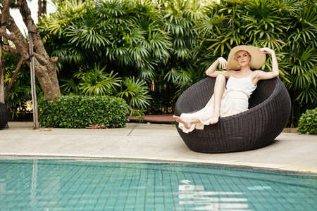 Elegant young adult Caucasian woman wearing stylish outfit with hat relaxing on wicker chair at poolside on summer vacation dayの写真素材