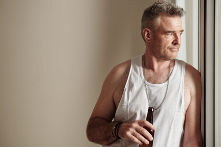 Portrait of pensive mature man with bottle of beer standing at window