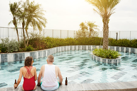 Middle-aged couple sitting by rooftop swimming pool, drinking beer and enjoying sunset