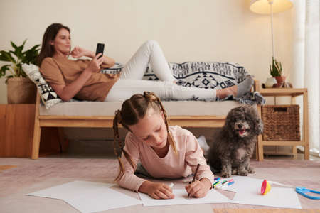 Girl lying on floor next to her dog and drawing pictures when mother reading in backgroundの写真素材