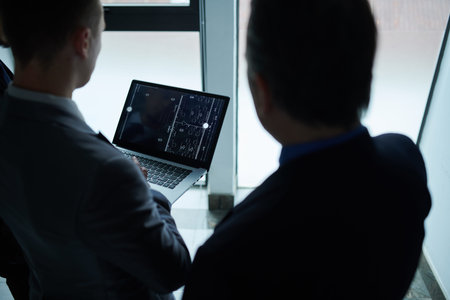 Business colleagues standing at office window and discussing building blueprint on laptop
