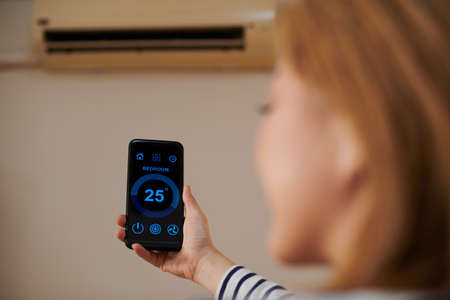 Woman using smart home application on smartphone to control air conditioner and set temperature in roomの写真素材