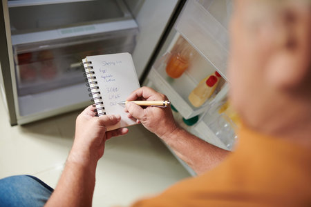 Elderly man checking his empty fridge and writing down shopping list in notepad