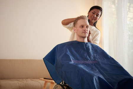 Smiling female caregiver cutting hair of young man with disability sitting in wheelchairの写真素材