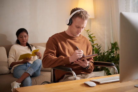 Young man with disability listening to music and working on tablet computer at home, girlfriend...