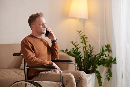 Serious young man with disability sitting in wheelchair and talking on phone