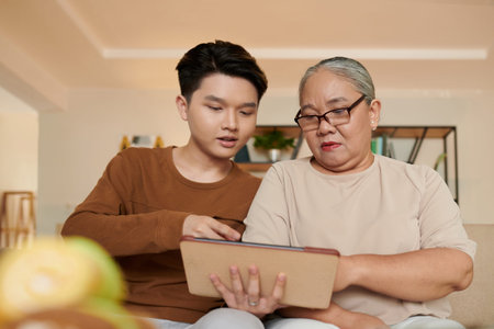 Vietnamese teenage boy showing grandmother how to use tablet computer for watching her favorite showsの写真素材