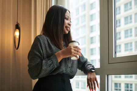 Portrait of smiling elegant female entrepreneur with cup of take out coffee standing at window in...