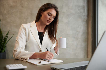 Portrait of businesswoman drinking cup of coffee and writing in plannerの写真素材