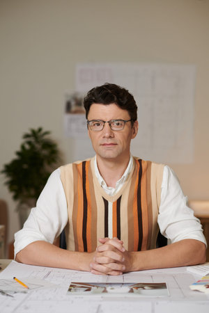 Portrait of serious confident architect in glasses sitting at desk with blueprints and looking at cameraの写真素材