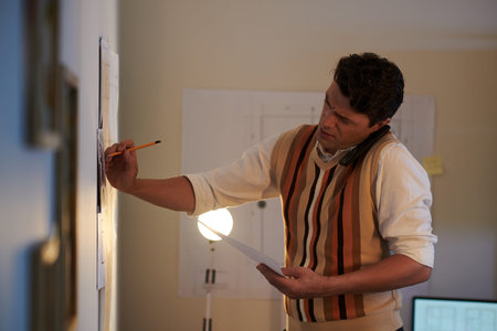 Frowning engineer talking on phone with client and correcting blueprint on wall