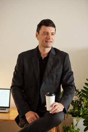 Portrait of smiling businessman sitting on office table with cup of take-out coffee in hand