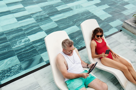 Rich middle-aged couple resting on chaise-lounges and reading news artciles online