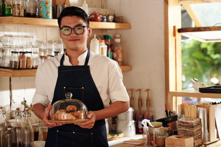 Portrait of smiling coffeeshop barista holding plate of freshly baked croissantsの写真素材