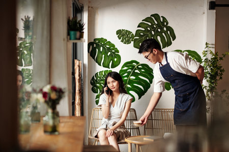 Coffeeshop waiter bringing cup of coffee to young woman talking on phoneの写真素材