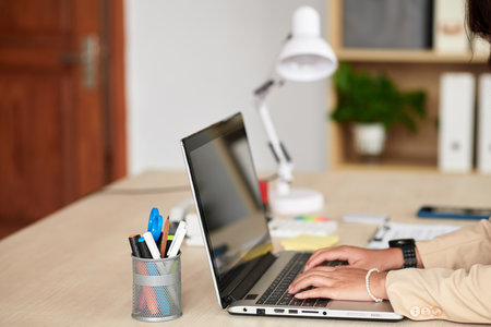 Close-up of young woman sitting at her table at office and typing on computer laptopの写真素材
