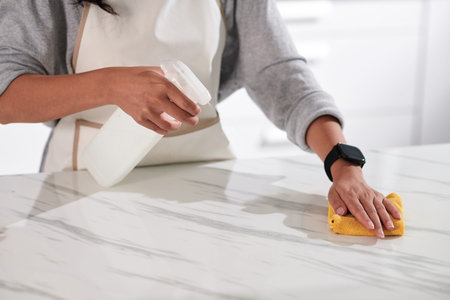 Close-up of housewife in apron cleaning table with rag and spray at home