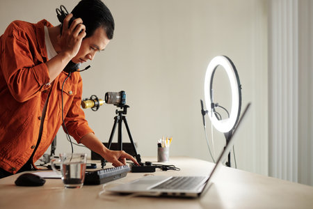 Side view selective focus of modern Asian influencer standing at desk in studio adjusting sound using mixing console and headphonesの写真素材