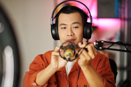 Medium close-up selective focus of Asian influencer wearing headphones sitting in studio getting ready for live streaming adjusting microphoneの写真素材
