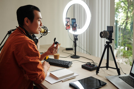Horizontal side view of modern Asian influencer sitting at desk making review of new wireless earbudsの写真素材