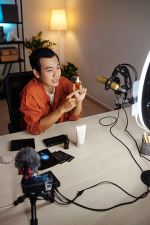 Vertical high angle view of young adult Asian male influencer making review of new beauty products demonstrating moisturizing serum or nourishing oil on cameraの写真素材