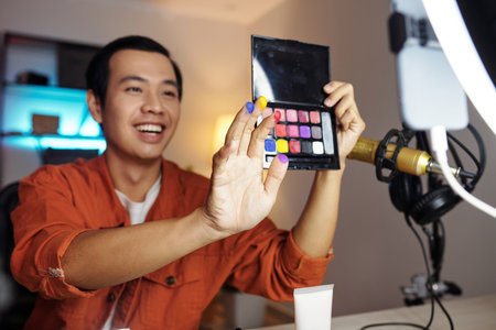 Horizontal selective focus medium shot of young Asian male influencer making video for blog demonstrating new cream eyeshadow palette colors on cameraの写真素材
